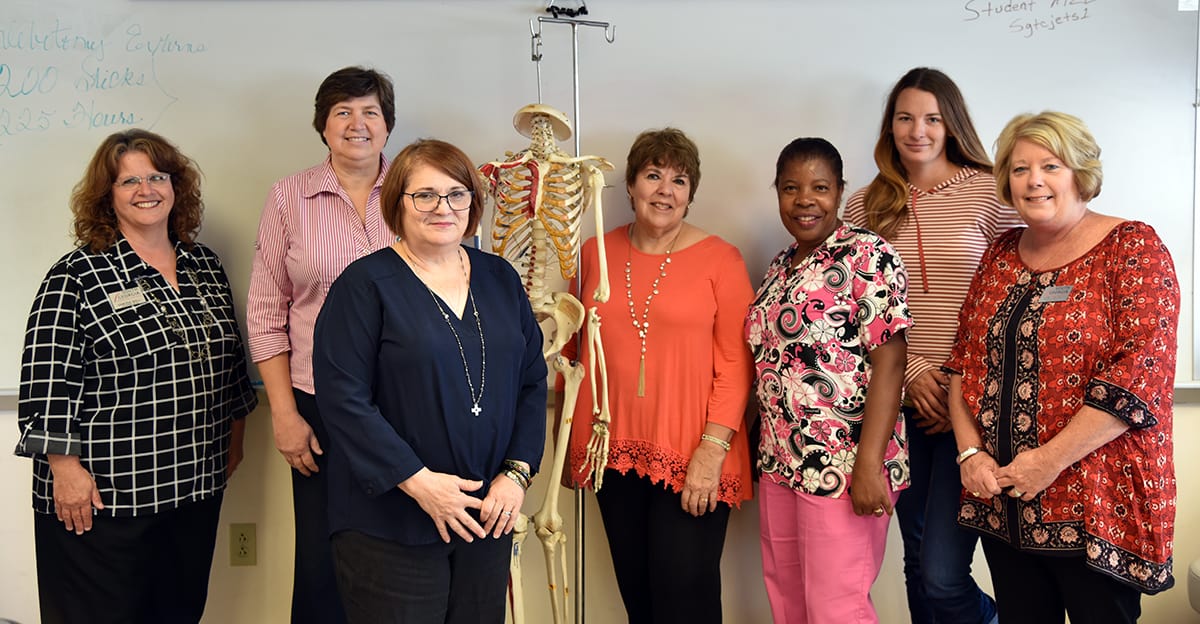The Medical Assisting Advisory Committee, a group of seven females, stand against a whiteboard in a classroom and pose for a photograph.