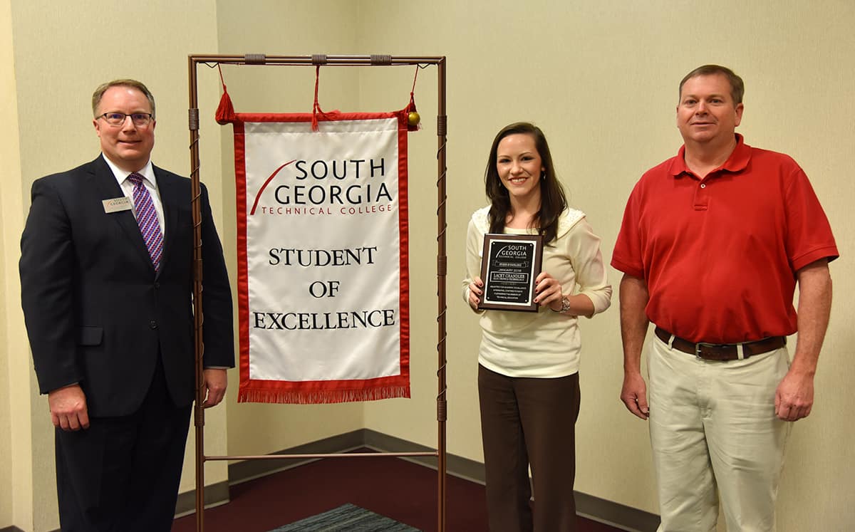 Student of Excellence winner Lacey Chandler (center) stands with nominating instructor Mike Collins (right) and SGTC Vice President of Academic Affairs David Kuipers (left) beside the Student of Excellence banner.