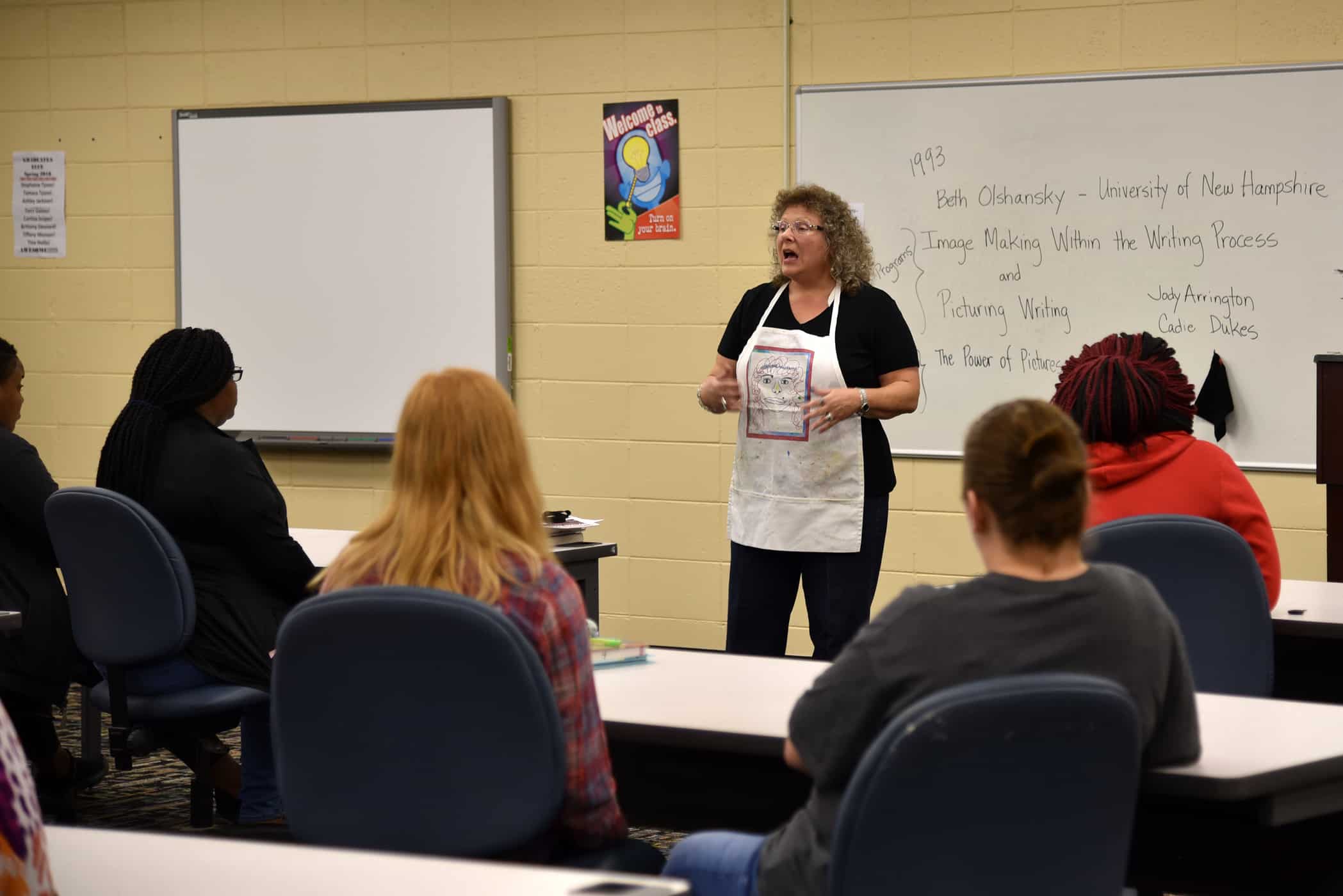 A woman in a white apron stands in front of a classroom.