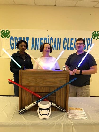 Three people stand behind a table, dressed in Star Wars costumes and holding light sabers.