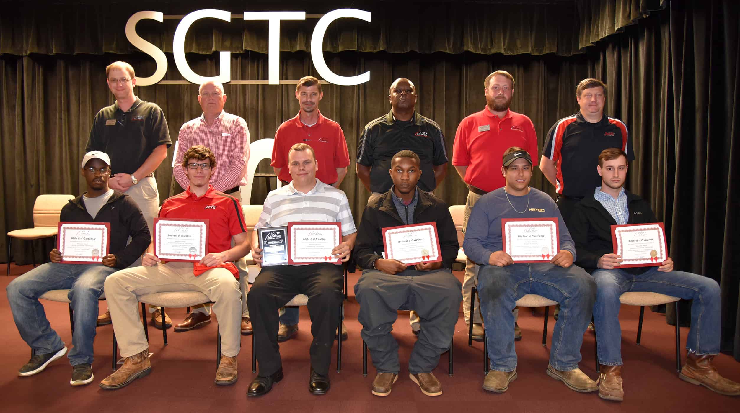 Six males sit in a row of chairs holding certificates. Behind them stand six males.