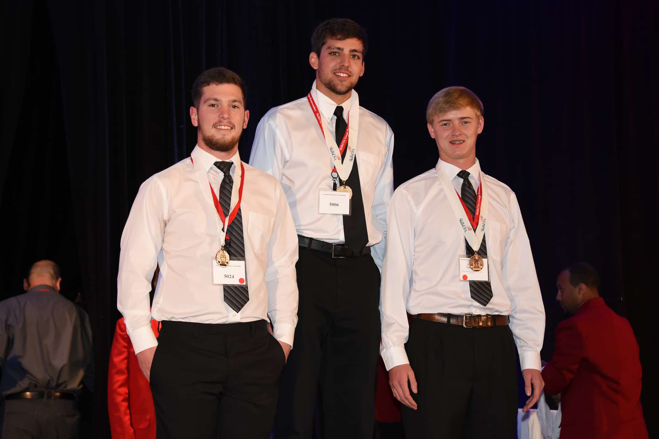 Three white males stand on top of a podium. The center male stands taller.