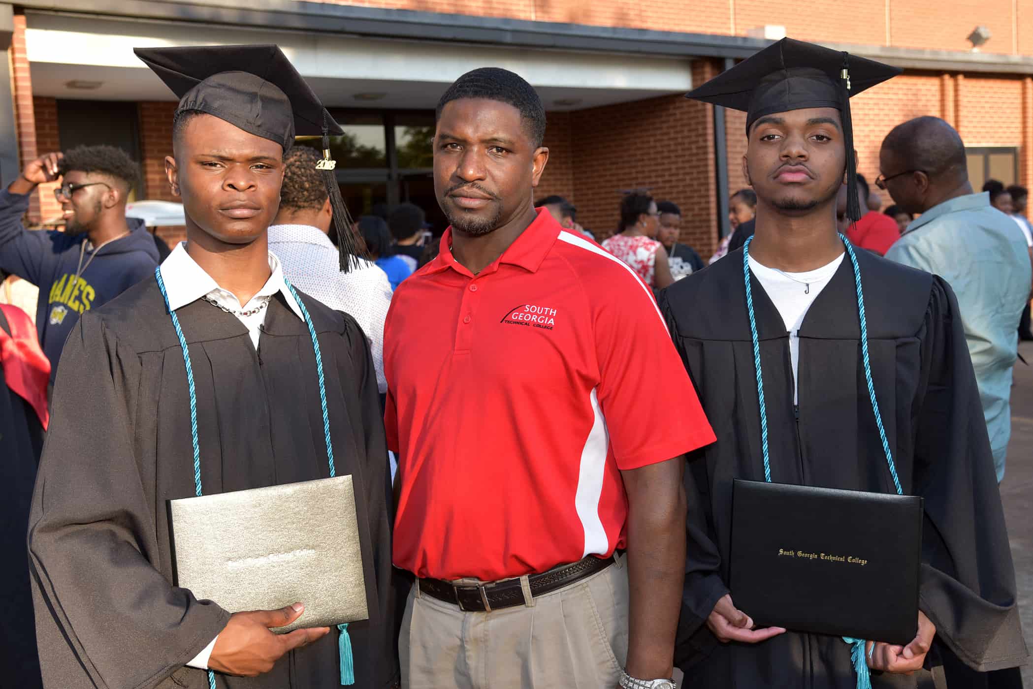 Two men in black graduation cap and gowns stand on either side of a man in a red shirt.